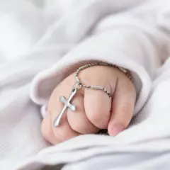 Dziecięca dłoń z krzyżykiem na bransoletce na tle białej tkaniny.