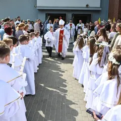 Dzieci w białych strojach komunijnych z zapalonymi świecami, biskup i księża przed kościołem.