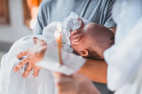 Dziecko w białej szacie trzymane na rękach, płonąca świeca.