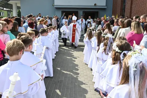 Dzieci w białych strojach komunijnych z zapalonymi świecami, biskup i księża przed kościołem.