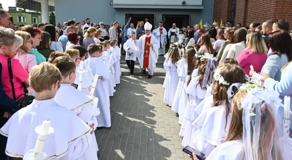 Dzieci w białych strojach komunijnych z zapalonymi świecami, biskup i księża przed kościołem.