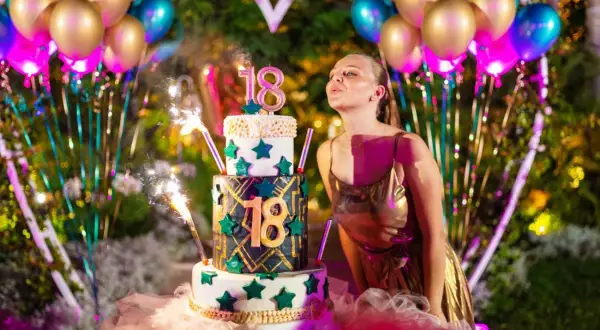 Dziewczyna w złotej sukience dmucha świeczki na torcie 18-stkowym, otoczona balonami i fajerwerkami.