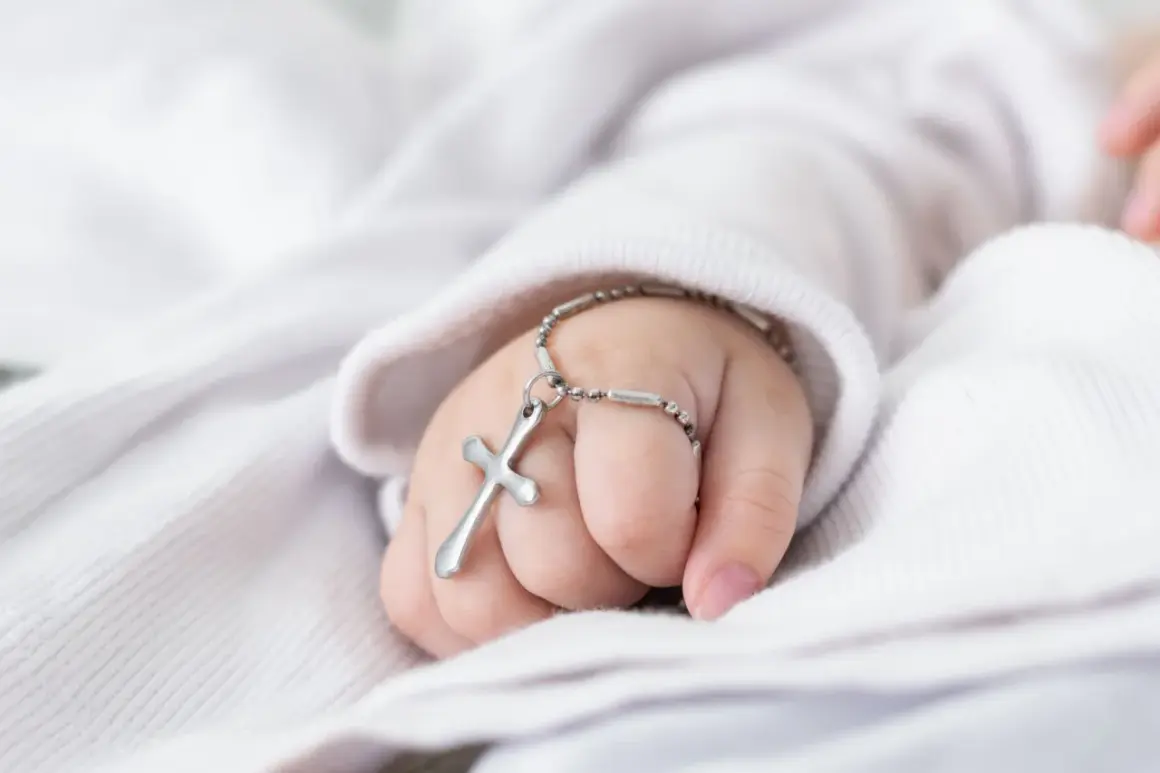 Dziecięca dłoń z krzyżykiem na bransoletce na tle białej tkaniny.