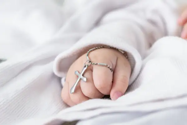 Dziecięca dłoń z krzyżykiem na bransoletce na tle białej tkaniny.